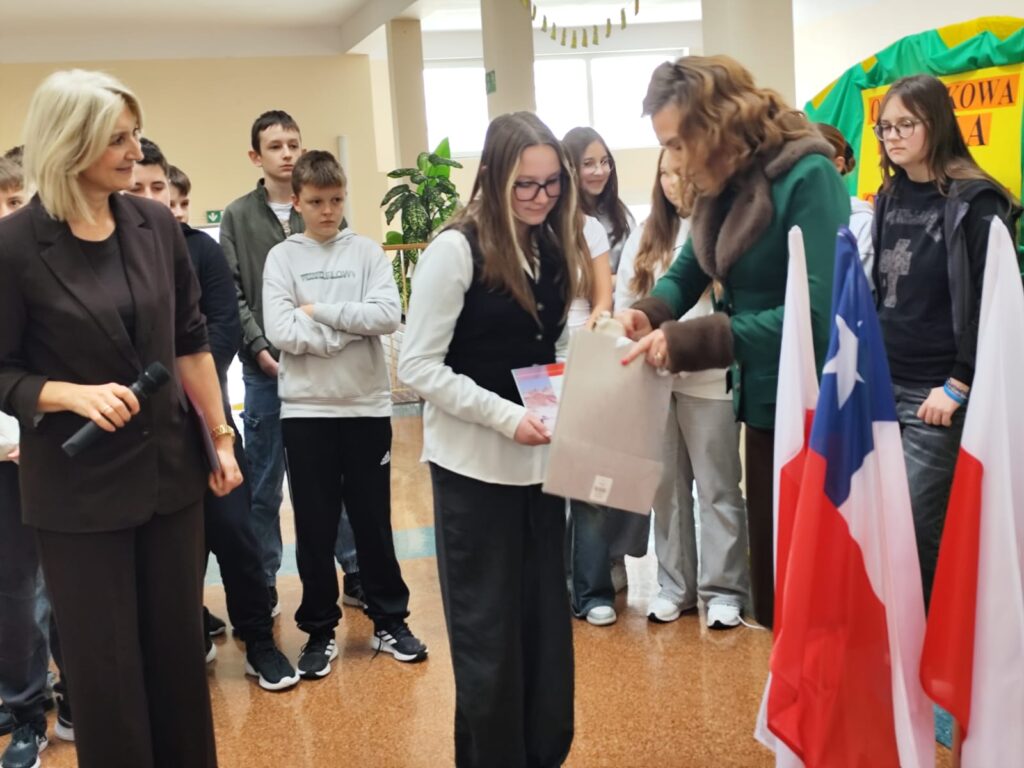 Uroczyste wręczenie biletów do Chile  na kolejną wymianę młodzieży. Na zdjęciu wiceprezes ds. prawnych Sierra Gorda SCM wręcza uczennicy bilet do Chile. W tle inni uczniowie. 