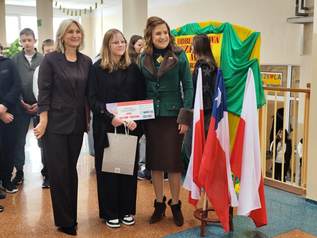 Uroczyste wręczenie biletów do Chile  na kolejną wymianę młodzieży. Na zdjęciu uczennica która otrzymała bilet do Chile na wymianę młodzieży. Po prawej stronie dwie flagi Polski i jedna Chile. 