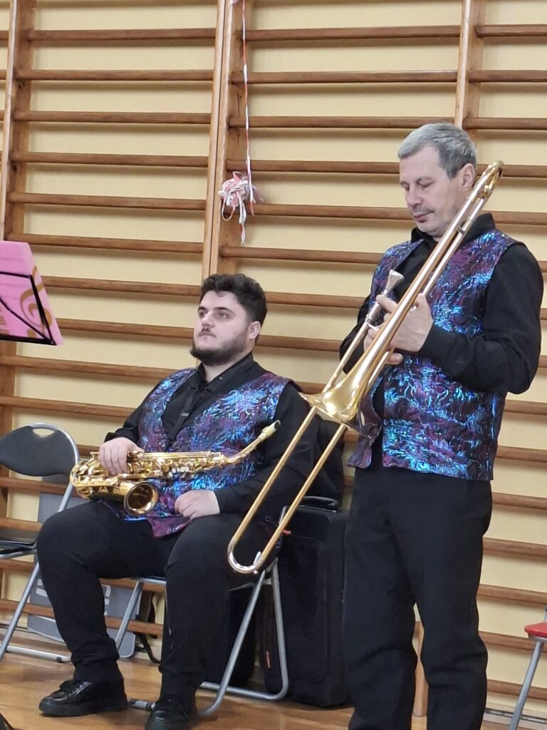 Koncert filharmonii. Na zdjęciu dwaj muzycy. Jeden z nich siedzi na krześle i trzyma saksofon, drugi stoi i trzyma w ręce puzon. 
