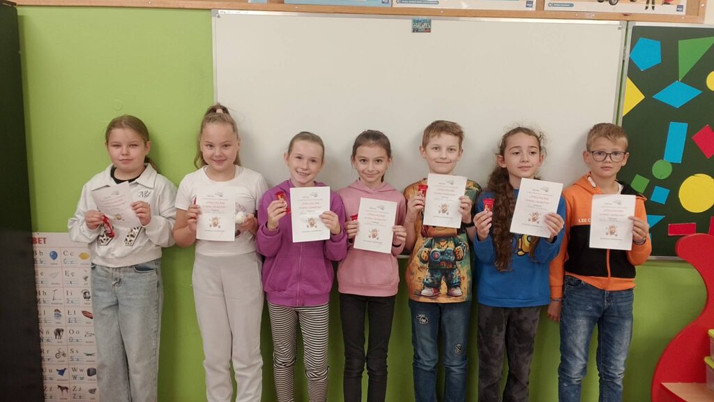 Szkolny Konkurs Literowania “Speeling Bee". Na zdjęciu uczniowie klasy trzeciej b, którzy brali udział w konkursie. Uczniowie stoją na tle białej tablicy, w rękach trzymają dyplom uczestnictwa w konkursie.