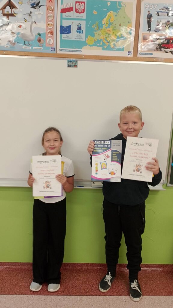 Szkolny Konkurs Literowania “Speeling Bee". Na zdjęciu zwycięzcy konkursu w klasie trzeciej b. Uczniowie stoją na tle białej tablicy, w rękach trzymają dyplom i nagrodę książkę.