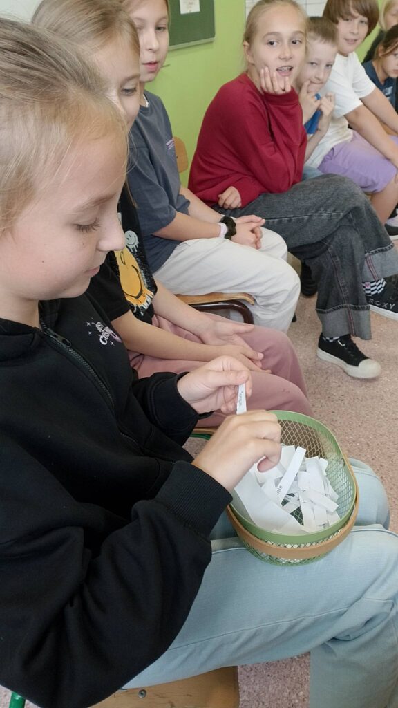 Szkolny Konkurs Literowania “Speeling Bee". Na zdjęciu uczniowie klasy trzeciej podczas konkursu. Na zdjęciu uczennica losuje słówko i literuje je w języku angielskim.