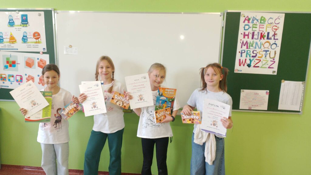 Szkolny Konkurs Literowania “Speeling Bee". Na zdjęciu uczennice klasy trzeciej a, które zajęły pierwsze i drugie miejsce w konkursie. Uczennice stoją na tle białej tablicy, w rękach trzymają dyplom i nagrodę książkę.