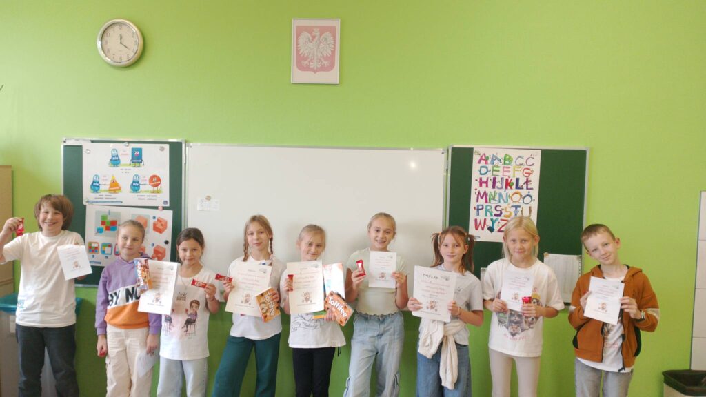 Szkolny Konkurs Literowania “Speeling Bee". Na zdjęciu uczniowie klasy trzeciej a, którzy brali udział w konkursie. Uczniowie stoją na tle białej tablicy, w rękach trzymają dyplomu uczestnictwa w konkursie.