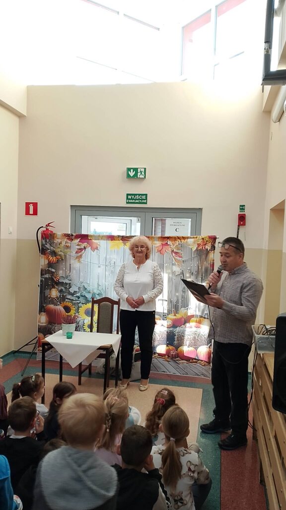 Na zdjęciu widać poetkę oraz nauczyciela, który trzyma mikrofon i przedstawia uczniom autorkę. Poetka stoi na tle banera o tematyce jesiennej. 