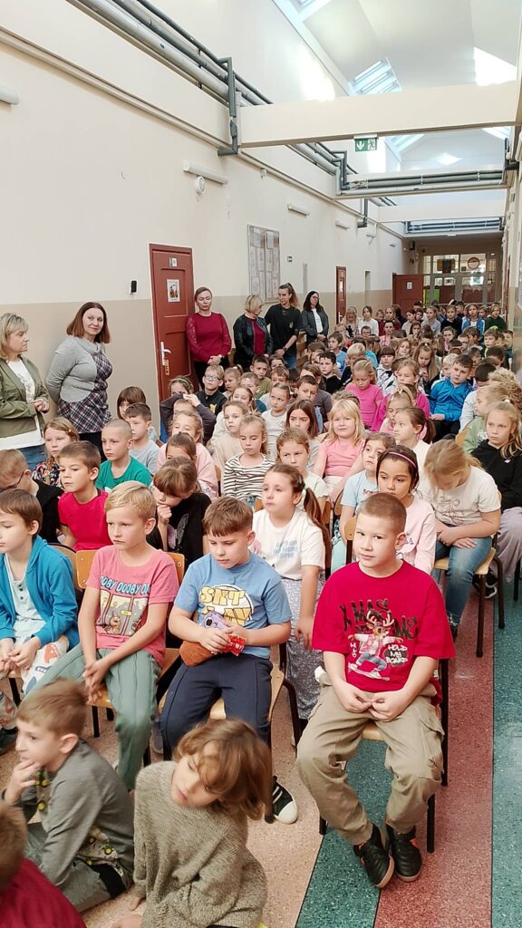 Na zdjęciu uczniowie klas 1-3 siedzą na krzesełkach na korytarzu i biorą udział w spotkaniu z pisarką wierszy i bajek dla dzieci.  