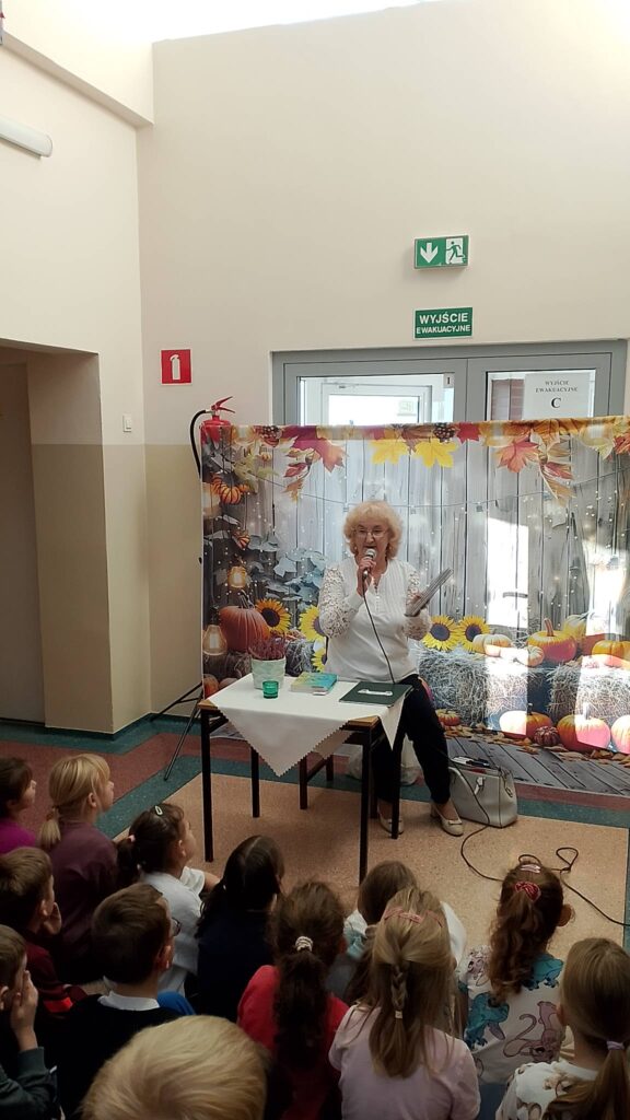 Zdjęcie przedstawia poetkę opowiadającą uczniom klas 1-3 historię powstania jej  wierszy. Na stoliku biała doniczka z wrzosem, w tle baner o tematyce jesiennej. Poetka siedzi na krześle przy stoliku, w lewej ręce trzyma książkę. 