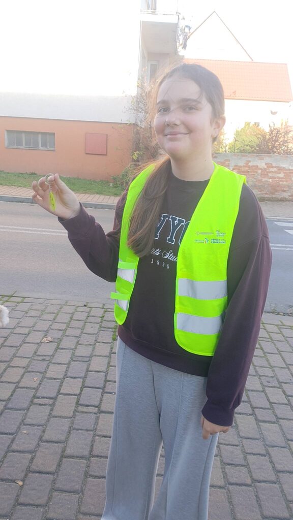 Odblaskowa Szkoła 2025- akcja rozdawania odblasków. Na zdjęciu uczennica ubrana w kamizelkę odblaskową z uśmiechem pokazuje odblask. 