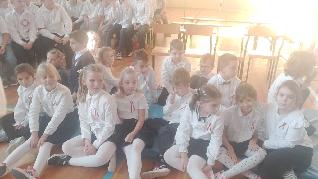 Apel z okazji Święta Niepodległości. Na zdjęciu uczniowie klasy pierwszej ubrani na galowo siedzą na materacu na sali gimnastycznej.