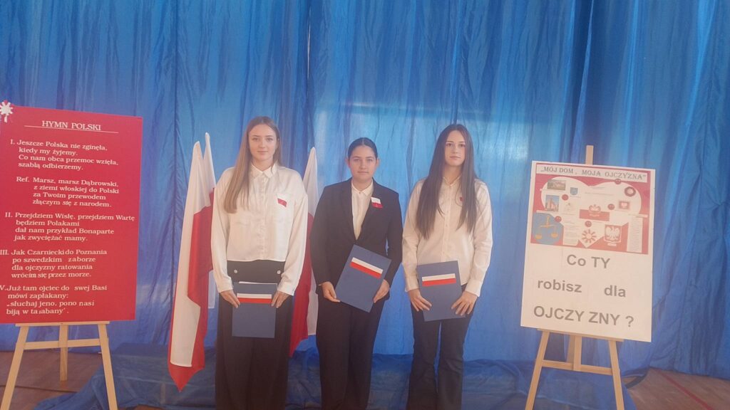 Apel z okazji Święta Niepodległości. Na zdjęciu uczennice klasy ósmej ubrane na galowo stoją na tle dekoracji. Po lewej stronie na czerwonym tle hymn Polski oraz plakat z napisem "Co ty robisz dla Ojczyzny?".