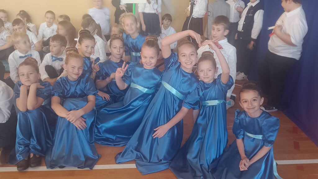 Apel z okazji Święta Niepodległości. Na zdjęciu uczennice klasy trzeciej ubrane w granatowe sukienki z uśmiechem pozują do zdjęcia, w tle chłopcu ubrani w białe koszule z niebieskimi muchami.