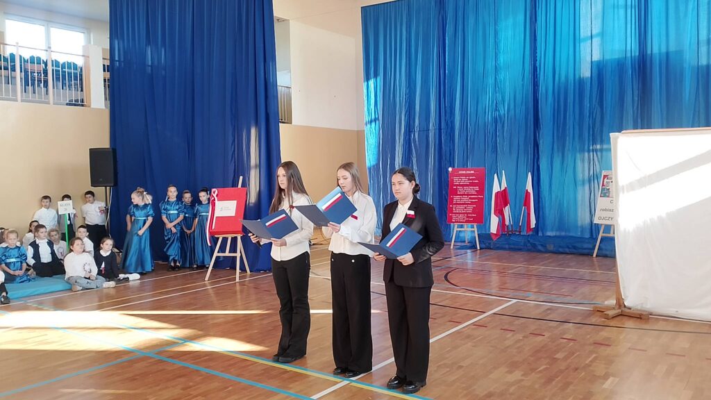Apel z okazji Święta Niepodległości. Na zdjęciu trzy uczennice klasy ósmej ubrane na galowo stoją na środku sali i prowadzą apel z okazji 11 listopada. W tle dekoracja patriotyczna.