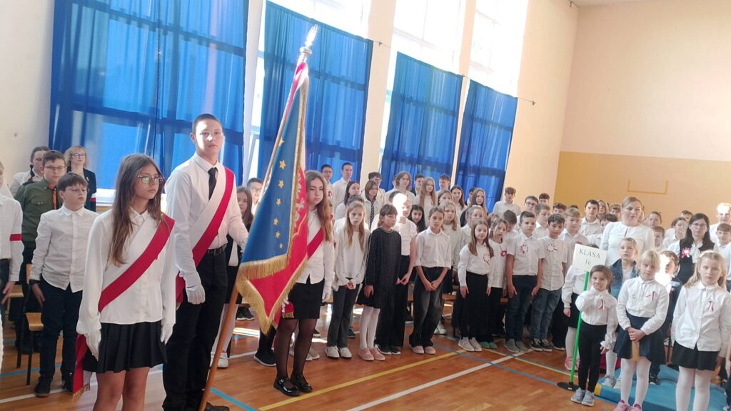 Apel z okazji Święta Niepodległości. Na zdjęciu poczet sztandarowy, chłopiec trzyma sztandar szkoły. Po prawej stronie społeczność szkolna ubrana na galowo.