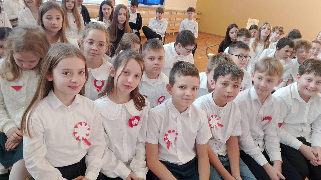 Apel z okazji Święta Niepodległości. Na zdjęciu uczniowie klasy piątej z uśmiechem pozują do zdjęcia, są ubrani na galowo, na ich białych koszulach widnieje biało-czerwony kotylion.
