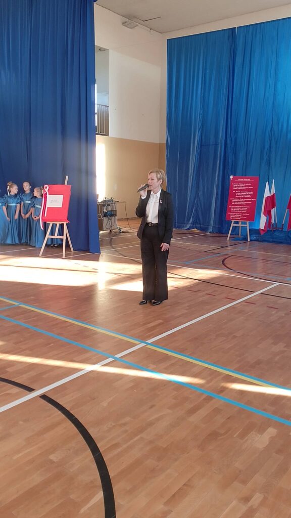 Apel z okazji Święta Niepodległości. Na zdjęciu Pani Dyrektor przemawia do uczniów, w tle dekoracja patriotyczna.