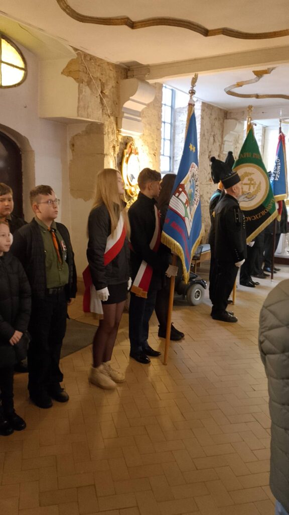 Udział sztandaru szkoły we Mszy Świętej w intencji Ojczyzny. Na zdjęciu poczet sztandarowy podczas mszy świętej. Chorąży trzyma sztandar, uczennice stoją po obu stronach chorążego. Wszyscy uczniowie mają biało- czerwone szarfy. 