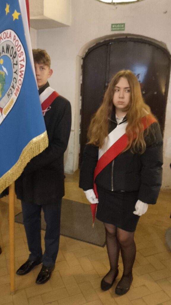 Udział sztandaru szkoły we Mszy Świętej w intencji Ojczyzny. Na zdjęciu uczennica z pocztu sztandarowego ubrana na galowo z biało-czerwoną szarfą. Obok stoi chorąży trzymający sztandar. 