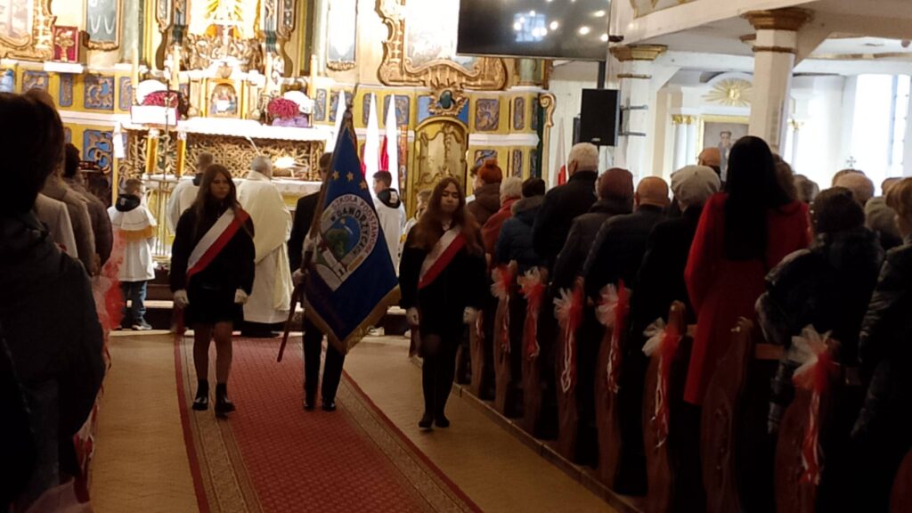 Udział sztandaru szkoły we Mszy Świętej w intencji Ojczyzny. Na zdjęciu poczet sztandarowy wychodzi z Kościoła, chorąży niesie sztandar, dwie uczennice idą po bokach. 