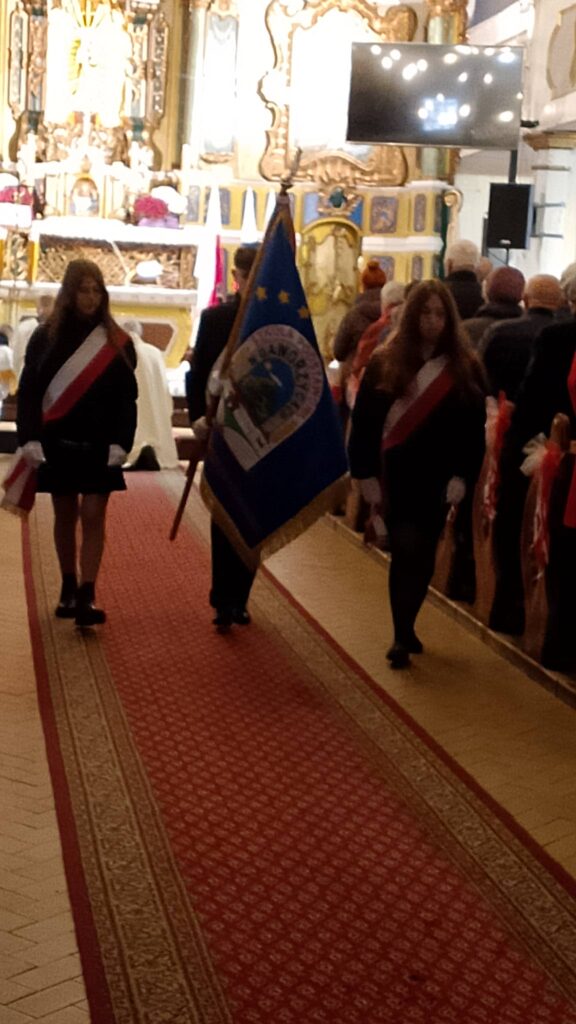 Udział sztandaru szkoły we Mszy Świętej w intencji Ojczyzny. Na zdjęciu poczet sztandarowy wychodzi z Kościoła, chorąży niesie sztandar, dwie uczennice idą po bokach. 