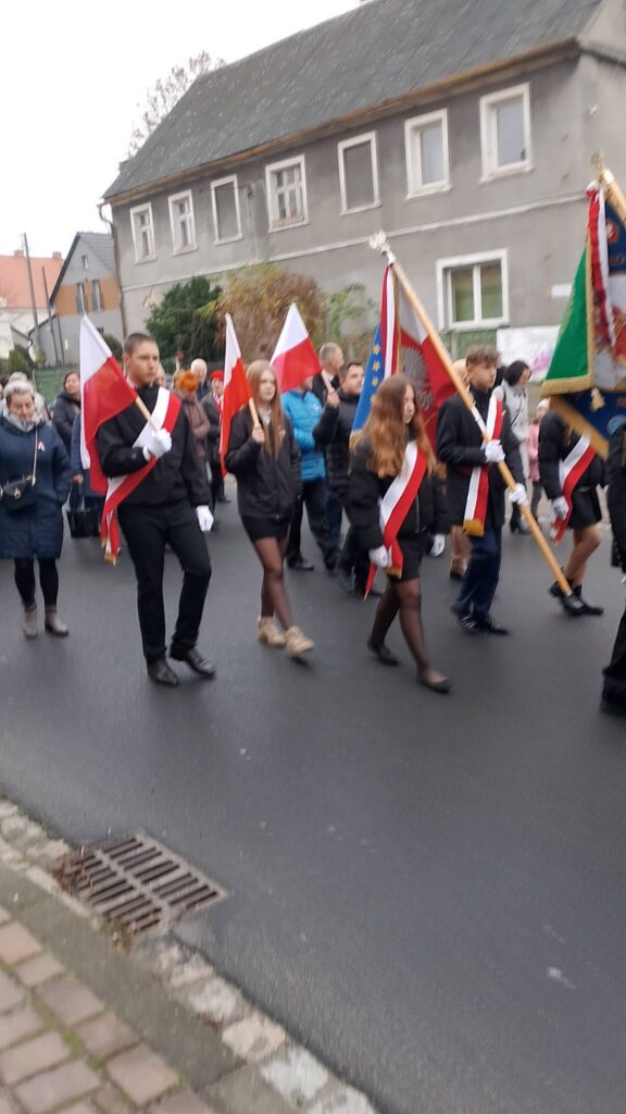 Udział sztandaru szkoły we Mszy Świętej w intencji Ojczyzny. Na zdjęciu poczet sztandarowy idzie w pochodzie, za sztandarem idą inni uczniowie niosąc flagi biało-czerwone.  