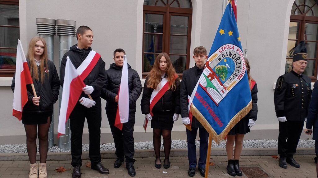 Udział sztandaru szkoły we Mszy Świętej w intencji Ojczyzny. Na zdjęciu poczet sztandarowy stoi pod budynkiem młyna, troje uczniów trzyma flagi, po prawej stronie uczeń stoi ze sztandarem. 