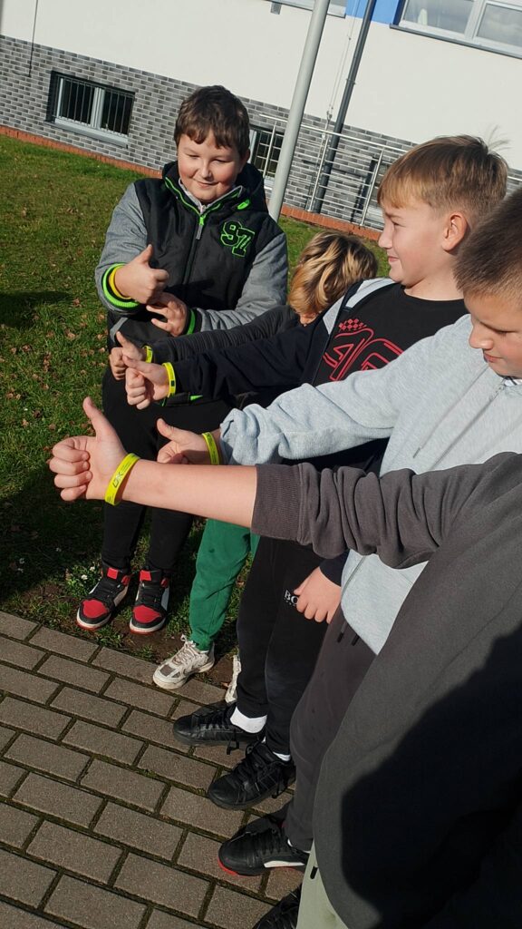 Odblaskowa Szkoła 2025- akcja rozdawania odblasków. Na zdjęciu chłopcy z klasy czwartej z wyciągniętymi rękami z założonymi opaskami odblaskowymi. 