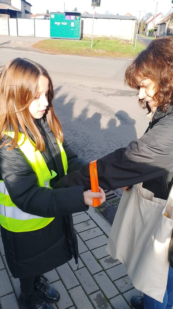 Odblaskowa Szkoła 2025- akcja rozdawania odblasków. Na zdjęciu uczennica zakłada kobiecie odblask na rękę. 