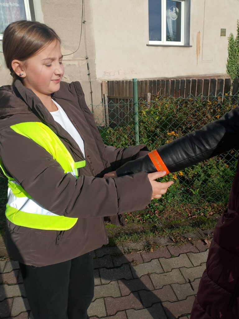 Odblaskowa Szkoła 2025- akcja rozdawania odblasków. Na zdjęciu uczennica zakłada kobiecie odblask na rękę. Uczennica uśmiecha się. 