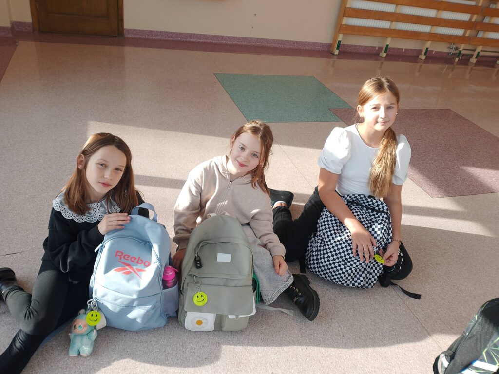 Odblaskowa Szkoła 2025- akcja rozdawania odblasków. Na zdjęciu uczennice klasy czwartej siedzą na korytarzu i prezentują swoje plecaki z zawieszonymi odblaskami. 