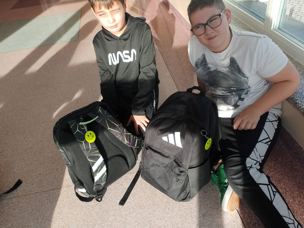 Odblaskowa Szkoła 2025- akcja rozdawania odblasków. Na zdjęciu uczniowie klasy czwartej siedzą na korytarzu i prezentują swoje plecaki z zawieszonymi odblaskami. 