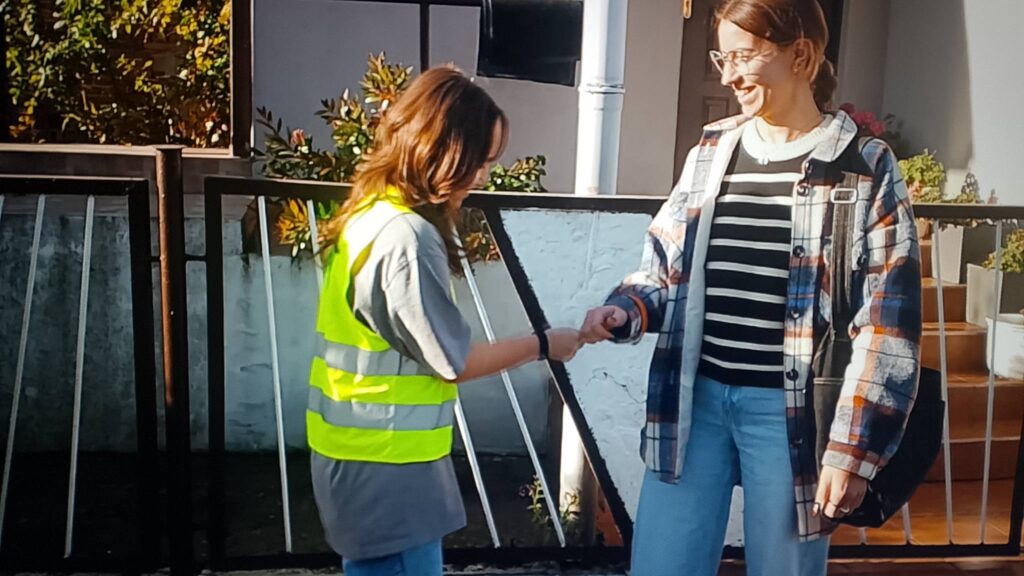 Odblaskowa Szkoła 2025- akcja rozdawania odblasków. Na zdjęciu uczennica zakłada kobiecie odblask na rękę. Uczennica i kobieta, która dostaje odblask  uśmiechają się. 