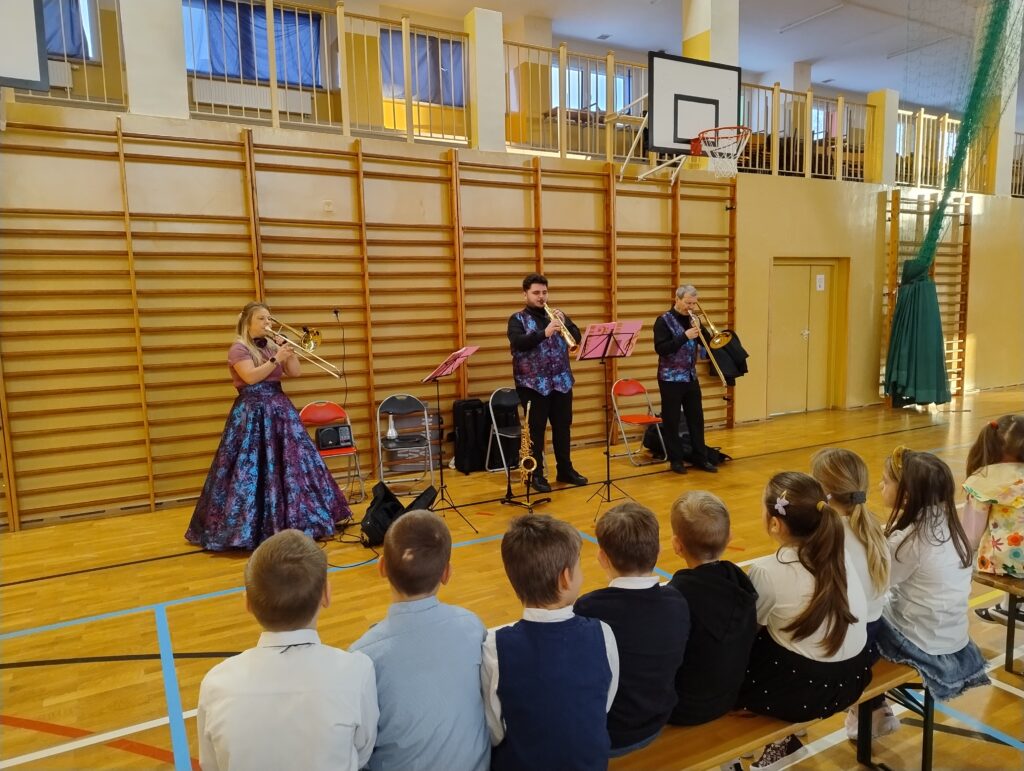 Koncert filharmonii. Na pierwszym planie zdjęcia widać dzieci siedzące w pierwszej ławce i oglądające koncert. Na drugim planie znajdują się muzycy filharmonii w trakcie grania na instrumentach. Kobieta w fioletowej sukni oraz mężczyzna po prawej stronie grają na puzonach, a mężczyzna stojący w środku gra na klarnecie sopranowym.