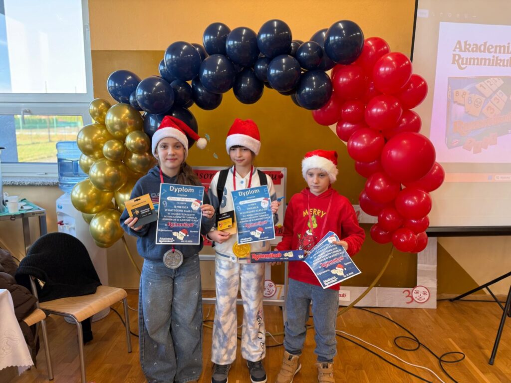 I Międzyszkolny Mikołajkowy Turniej Rummikub. Na zdjęciu uczniowie trzymają ręce dyplom i nagrody. Za nimi dekoeacja: balony złote, granatowe i czerwone.