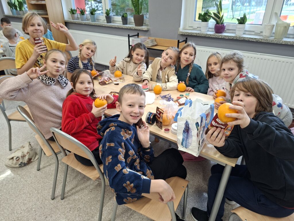 Warsztaty świąteczne w szkolnej bibliotece. Na zdjęciu uczniowie klasy trzeciej siedzą przy stoliku, każda osoba ma pomarańczę. Dodatkowo uczniowie trzymają suszone goździki. 