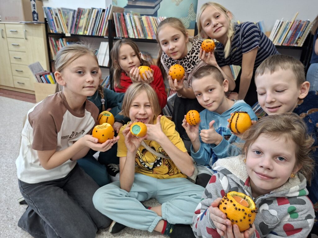 Warsztaty świąteczne w szkolnej bibliotece. Na zdjęciu uczniowie trzymają w ręce wykonaną ozdobę z pomarańczy, uczniowie są uśmiechnięci. 