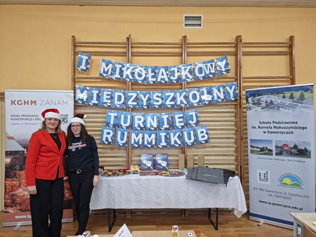 Przedstawia organizatorki ubrane w czerwoną marynarkę i bluzkę z napisem Trener Rummikub, które na głowach mają czerwone mikołajkowe czapki. Na drabinkach wisi biały napis I MIĘDZYSZKOLNY MIKOŁAJKOWY TURNIEJ RUMMIKUB. Po lewej stronie jest baner sponsora turnieju KGHM ZANAM, po prawej stronie jest szkolny baner ze zdjęciem Szkoły Podstawowej im. Kornela Makuszyńskiego w Gaworzycach. Na środku zdjęcia stoi stół nakryty białym obrusem z nagrodami: karty podarunkowe do Empiku, gra Rummikub, medale, breloczki z literą "R" (Rummikub) oraz bidony, torby i worki z logo KGHM ZANAM.