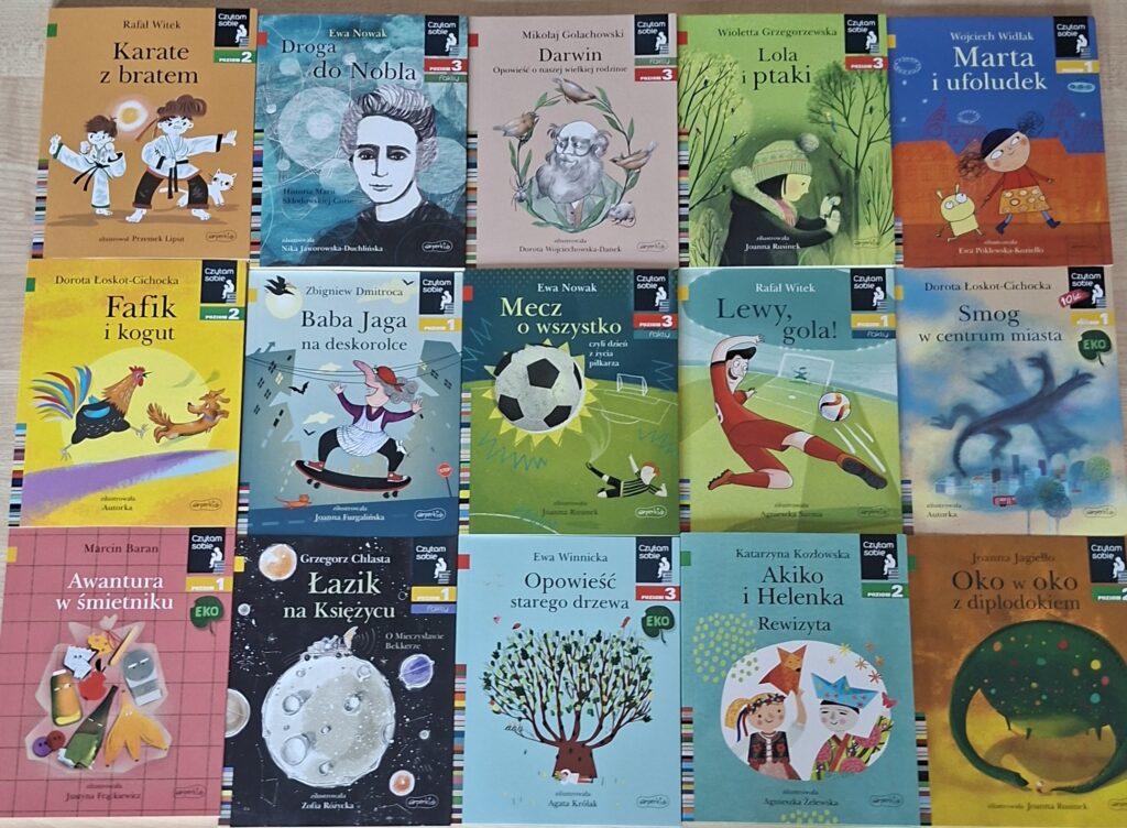 Przedstawia książki ułożone na stole obok siebie. Kolorowe Ilustracje okładek przedstawiają: koguta z psem, piłkarza z piłką, księżyc w kosmosie oraz ufoludka z dziewczynką.