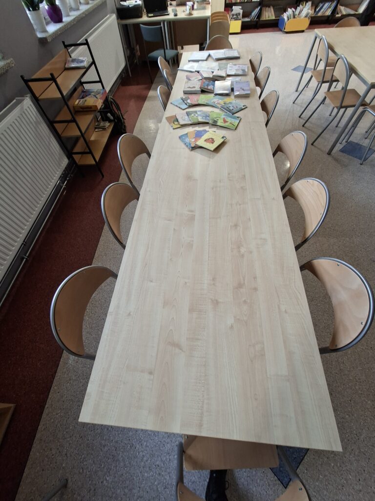 Przedstawia nowe stoły z krzesłami, wśród których znajdują się regały z książkami. Na stołach ułożone są książki. Stoły są jasnego koloru.