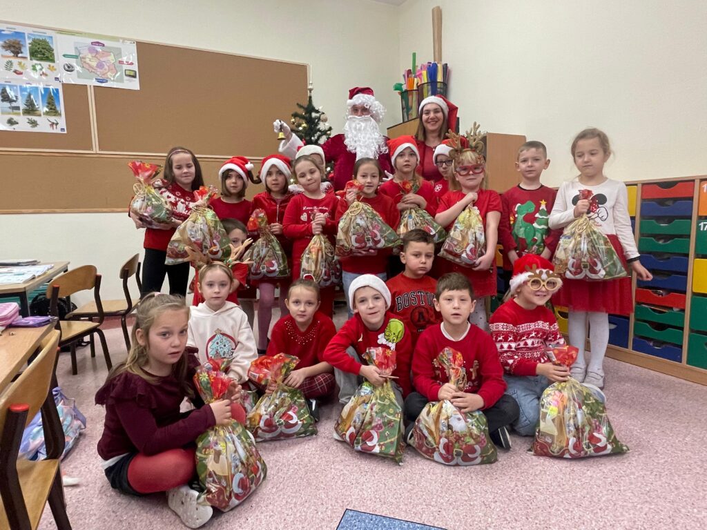 Mikołajki 2025. Na zdjęciu uczniowie klasy pierwszej pozują do zdjęcia ubrani mikołajkowo, za uczniami stoi choinka i Mikołaj trzymający w prawej ręce dzwoneczek. 