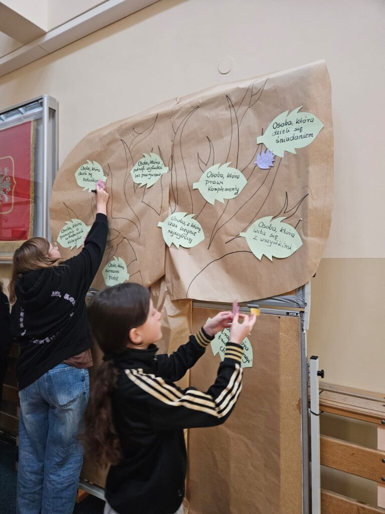 Na zdjęciu dwie uczennice stojące przed korkową tablicą. Dziewczyny stoją po lewej stronie. Przypinają zielone listki  z papieru do szkicu drzewa zrobionego z brązowego papieru na tablicy.
