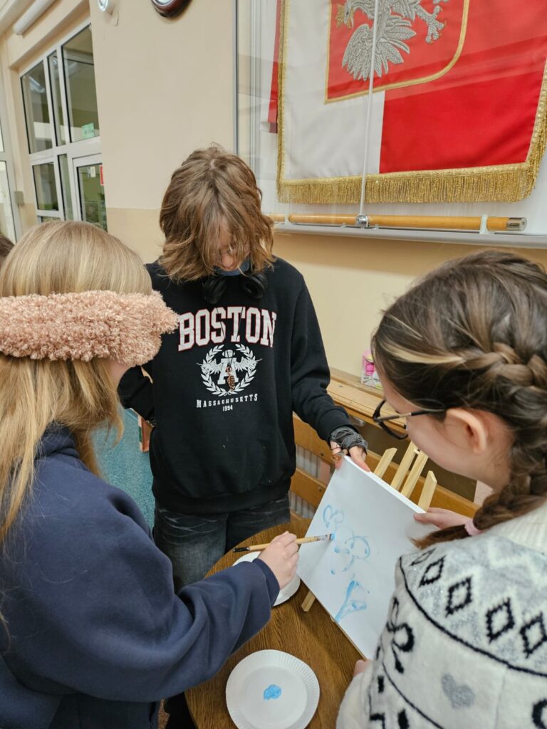 Tydzień Tolerancji i Szacunku 2025. Na zdjęciu trzy uczennice stojące przy drewnianym, okrągłym stoliku, stojącym na szkolnym korytarzu. Pierwsza uczennica od lewej ma zasłonięte oczy i w prawej ręce ma pędzelek do malowania .Maluje coś  w kolorze niebieskim na białym płótnie. Na środku stoi uczennica w czarnej bluzie, która trzyma białe płótno, aby ułatwić malowanie. Z prawej strony znajduje się uczennica, która stoi tyłem. Ona również podtrzymuje płótno, aby ułatwić malowanie obrazu.