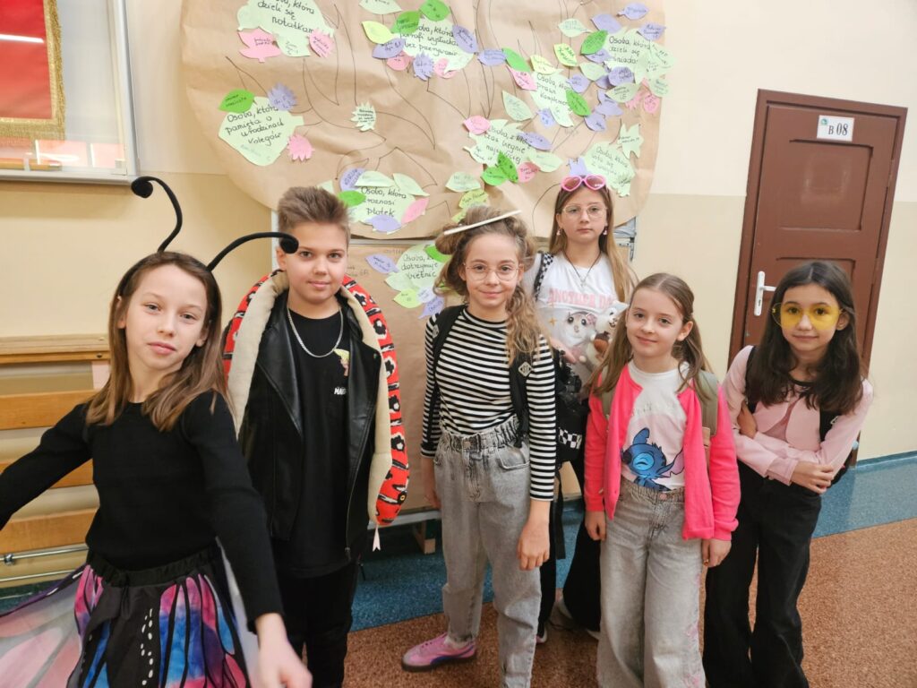 Na zdjęciu znajduje się sześcioro  uśmiechniętych uczniów stojących na korytarzu szkolnym. Stoją przed tablicą z drzewem Życzliwych Ludzi. Od lewej: dziewczynka przebrana za motylka, drugi jest chłopiec w skórzanej czarnej kurtce z maskotką węża na szyi, trzecia to dziewczynka w koszulce w paski , jeansach i fikuśniej fryzurze. Kolejna dziewczynka ma na głowie różowe okulary, na nosie zwykłe okulary a w ręku maskotkę .Następna dziewczynka ma dwa kitki, jaskrawy kardigan, białą koszulkę i szerokie niebieskie jeansy. Ostatnią dziewczynka ma długie ciemne włosy. Na nosie ma żółte okulary. Ubrana jest w różowy sweter i czarne spodnie.