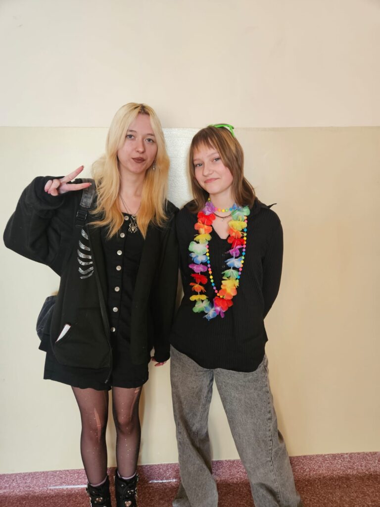 Na zdjęciu dwie uczennice stojące przy ścianie na korytarzu szkolnym. Po lewej stronie dziewczyna o blond włosach, ubrana w czarną bluzę, czarne spodenki oraz rajstopy z dziurami. Obok stoi niższa dziewczyna o krótszych i ciemniejszych włosach. Na głowie ma zielone okulary, na szyi wianek z kwiatów w stylu hawajskim i kolorowe korale. Dziewczyna ubrana jest w czarną bluzę oraz szerokie , szare jeansy.