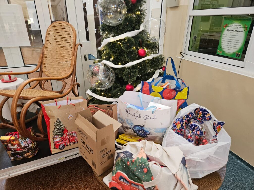 Na zdjęciu korytarz szkolny. Na pierwszym planie po prawej stolik z torbami i reklamówkami wypełnionymi słodyczami. Z lewej strony znajduje się podest, na którym stoi fotel bujany, przy którym znajdują się kolejne torby. Z tyłu stoi wysoka sztuczna, udekorowana choinka.