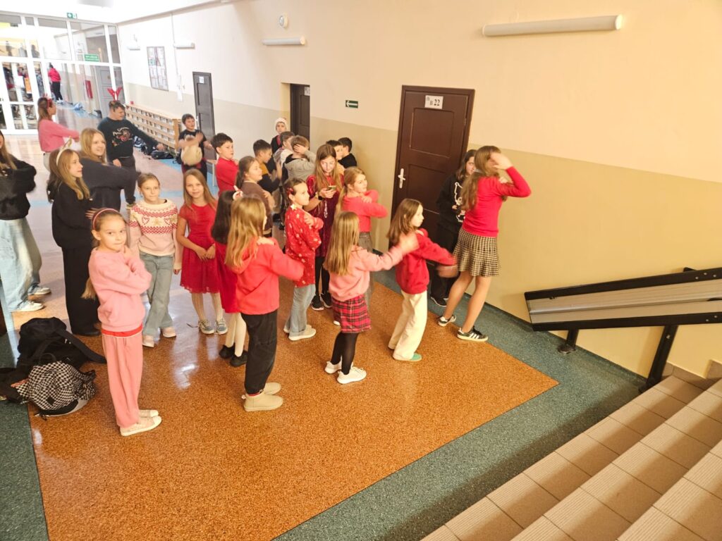 Na zdjęciu fragment schodów i korytarz na segmencie A. Na korytarzu stoi grupa kilkunastoosobowa ubrana przeważnie w kolorze czerwieni. Uczniowie w większości skierowani są w jednym kierunku i wykonują takie same ruchy.