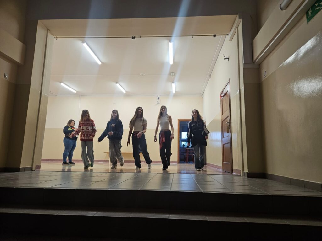 Na zdjęciu  oświetlony korytarz szkolny. Na środku znajdują się uczennice ubrane w stroje sportowe. Pokazują kroki do tańca.