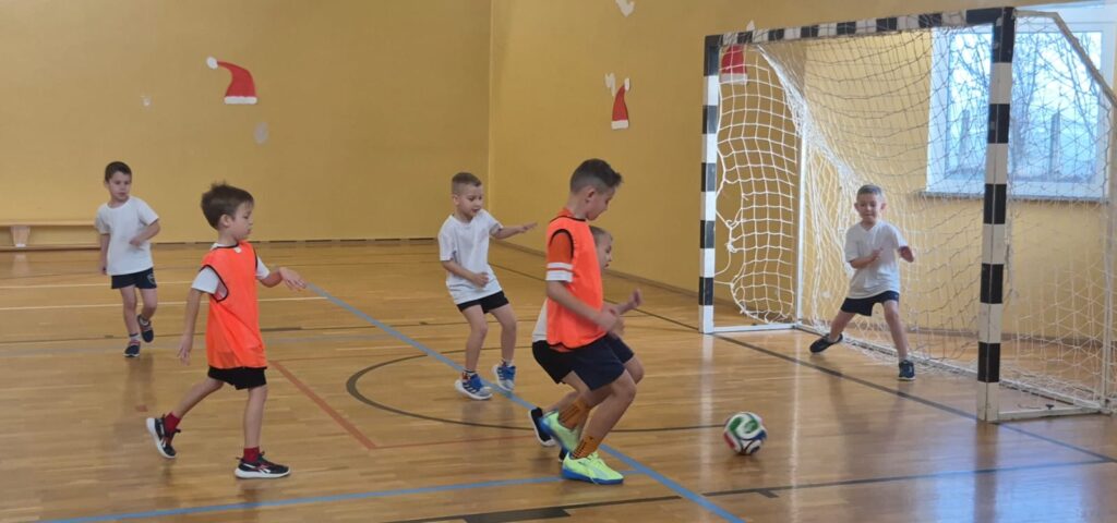 Turniej mikołajkowy. Na zdjęciu uczniowie klasy pierwszej grają w piłkę nożną na sali gimnastycznej. Dwóch uczniów ma na sobie pomarańczowe kamizelki.