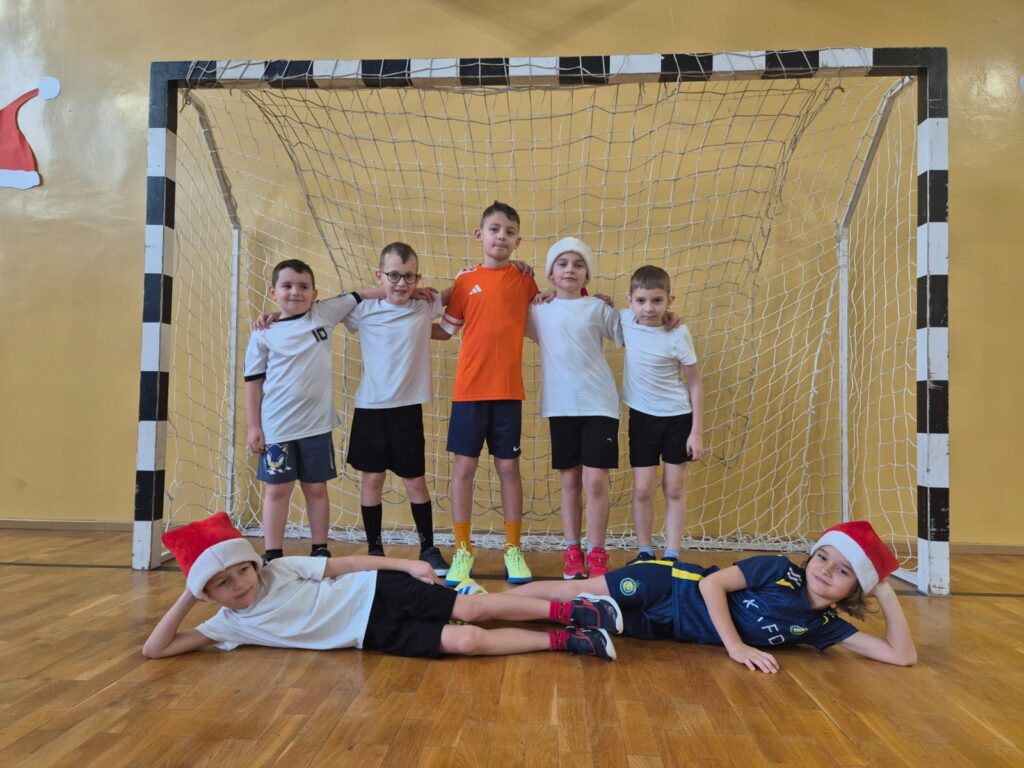 Turniej mikołajkowy. Na zdjęciu sześciu uczniów pozuje na tle bramki do zdjęcia, dwóch chłopców leż na przeciwko siebie na podłodze.
