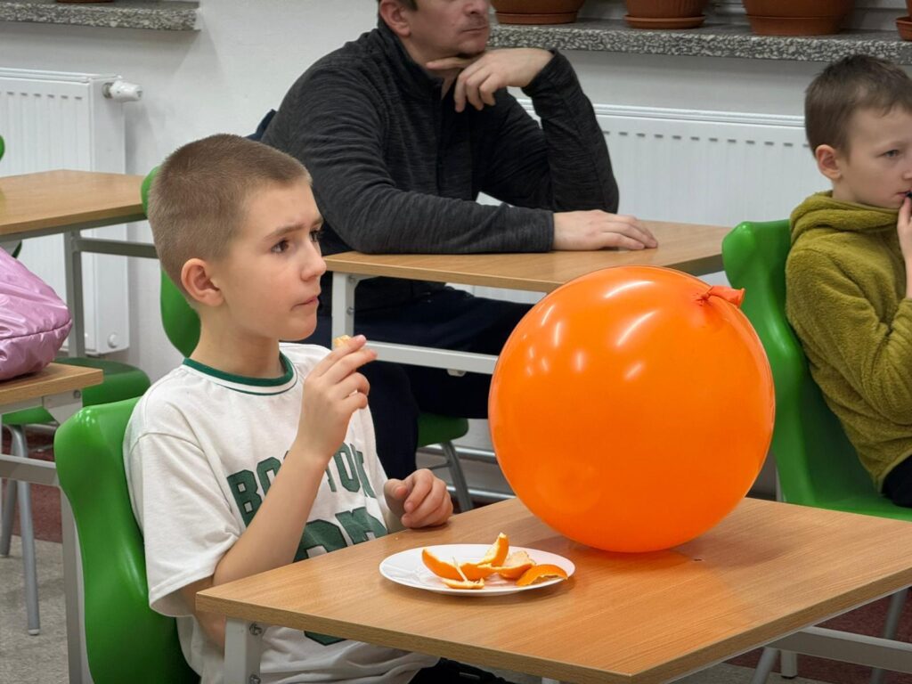 Wywiadówka z Kultury Fizycznej. Na zdjęciu uczeń klasy drugiej je pomarańczę. Na jego ławce leżu pomarańczowy balon.