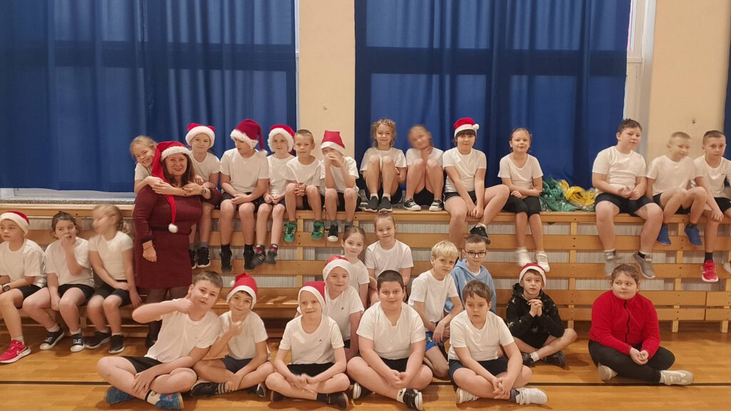 Turniej mikołajkowy. Na zdjęciu uczniowie klasy drugiej. Część uczniów siedzi na podłodze, część stoi, niektórzy mają mikołajkowe czapki.